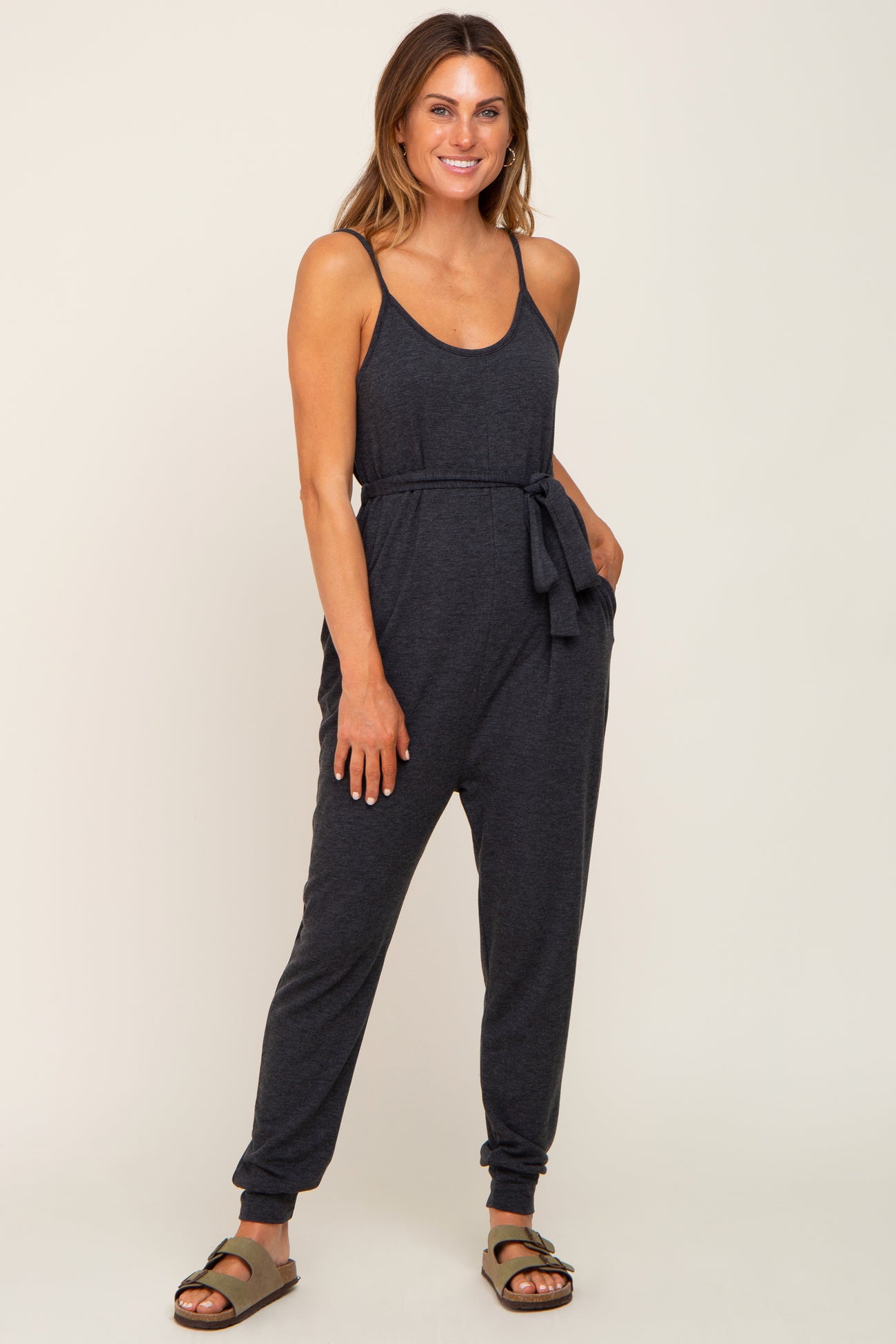 woman wearing a dark gray sleeveless jumpsuit with a tie waist and tapered legs, styled with sandals