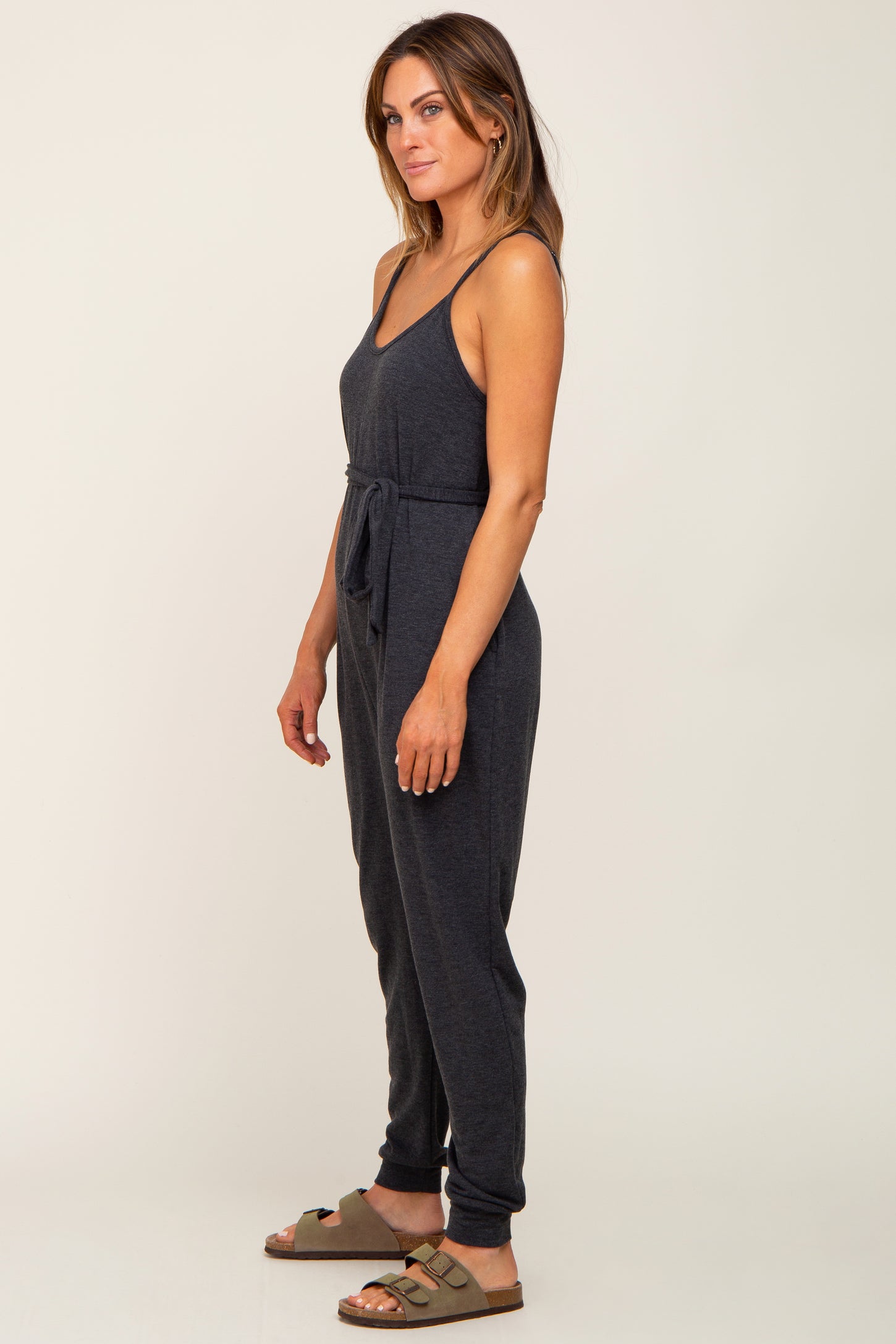 woman wearing a charcoal grey sleeveless maternity jumpsuit with a waist tie and olive green sandals