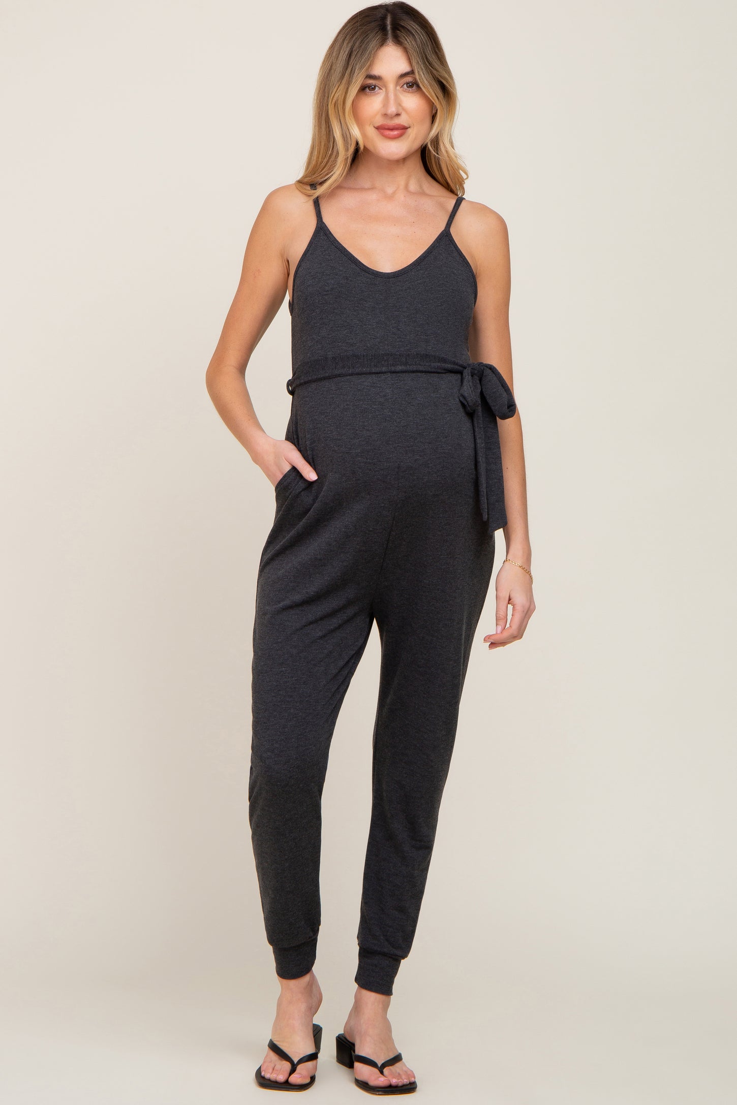 woman wearing a dark gray maternity jumpsuit with a tie waist and pockets, showcasing her baby bump