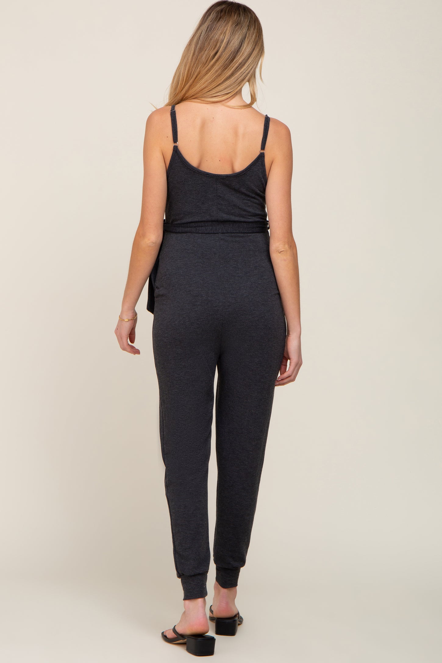 woman wearing a dark gray maternity jumpsuit, showing the back view with tapered legs and adjustable straps