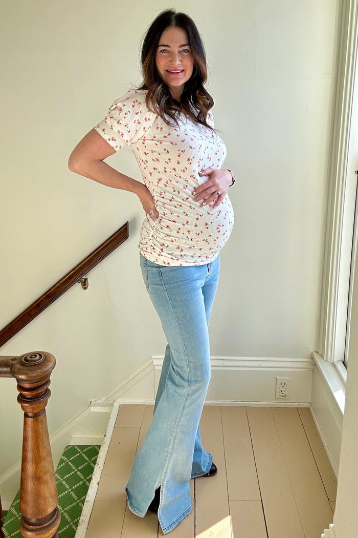 woman wearing a floral print maternity top and blue flared jeans, posing with her baby bump visible