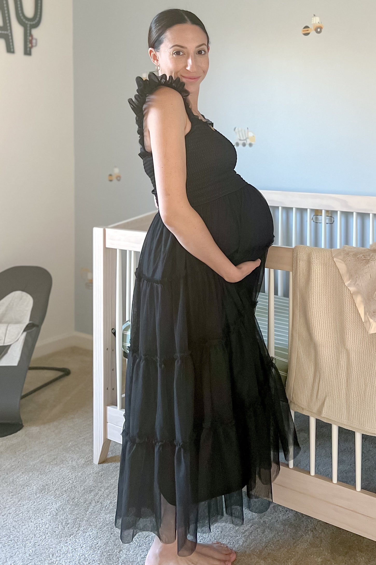 woman wearing a black tiered maternity dress with ruffled sleeves and a visible baby bump while standing next to a crib
