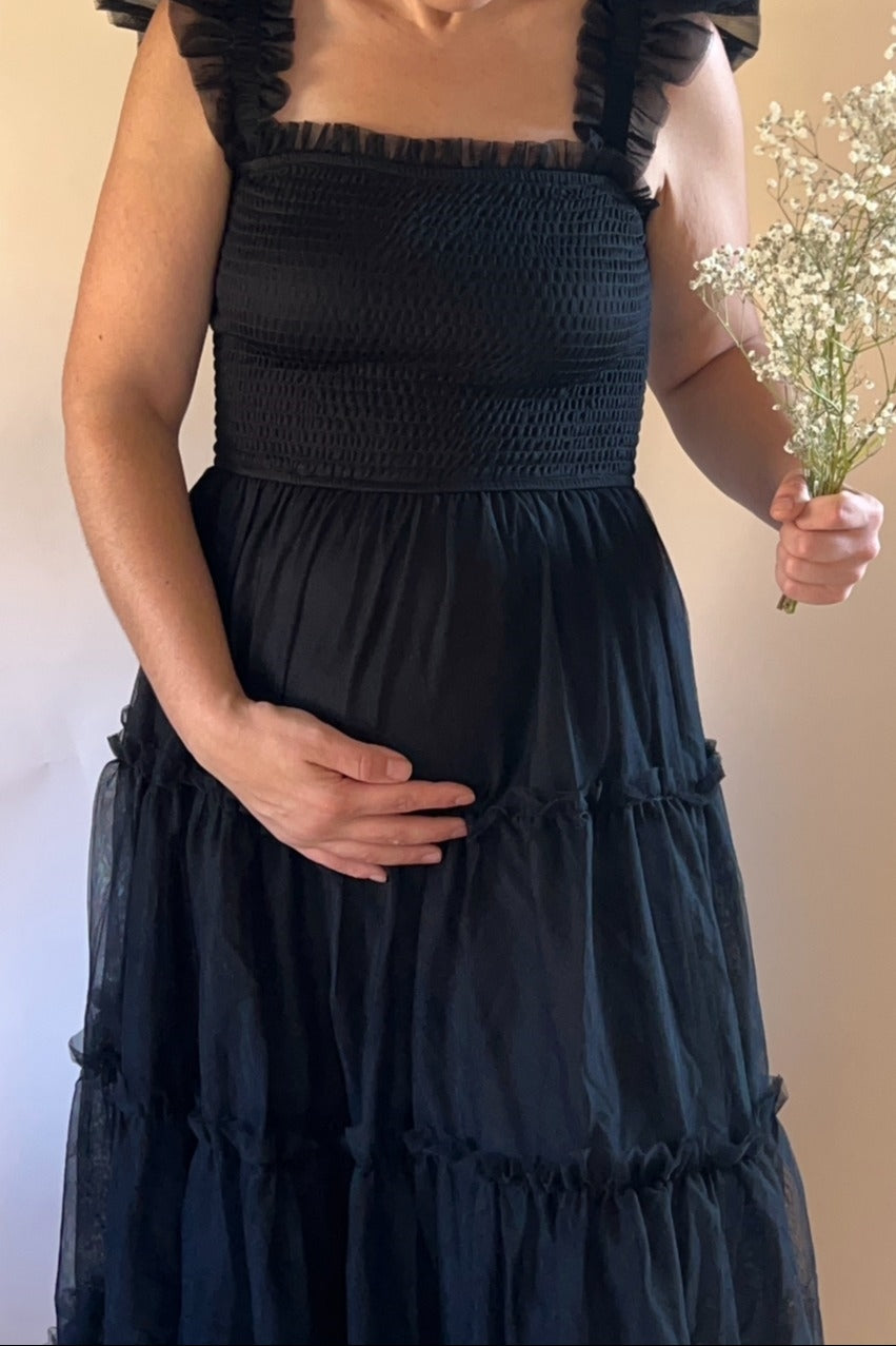 woman wearing a black smocked maternity dress with ruffled straps and holding a baby's breath flower while showing her baby bump