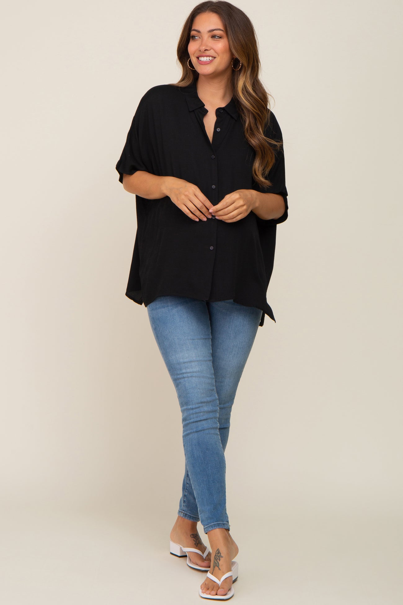 woman wearing a black maternity top with buttons, paired with blue skinny jeans and white sandals, standing confidently