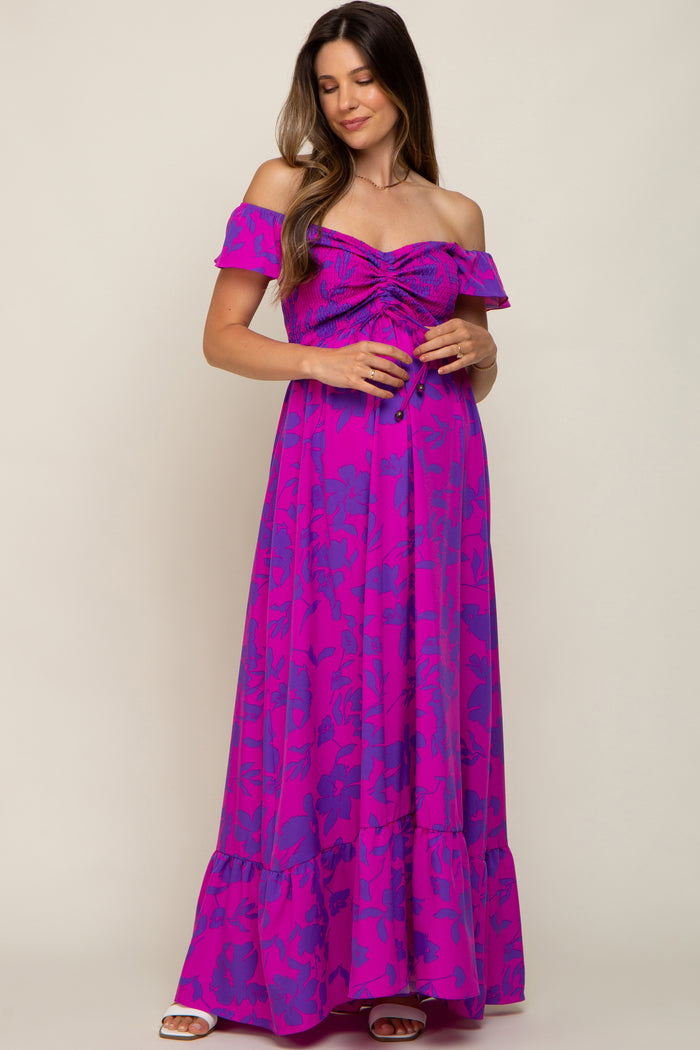 woman wearing a purple off-shoulder maternity maxi dress with gathered detailing and a flowy skirt, holding her baby bump