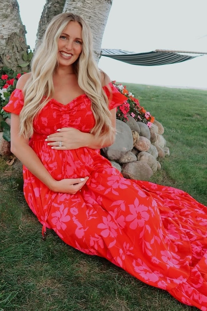 woman wearing a red floral maternity dress sitting on the grass and cradling her baby bump