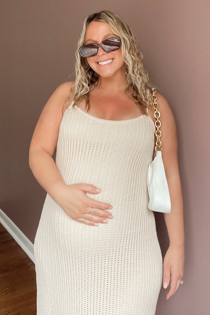 woman wearing a beige knitted maternity dress and holding her baby bump, accessorized with sunglasses and a white handbag