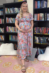 Smiling pregnant woman in a floral maxi dress standing in front of bookshelves in a library or classroom.