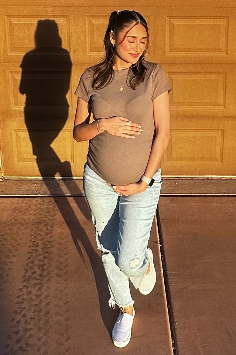 woman wearing a brown maternity t-shirt and light-wash jeans, holding her baby bump, standing in front of a garage door