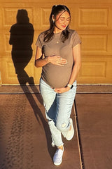 woman wearing a brown maternity t-shirt and light-wash jeans, holding her baby bump, standing in front of a garage door