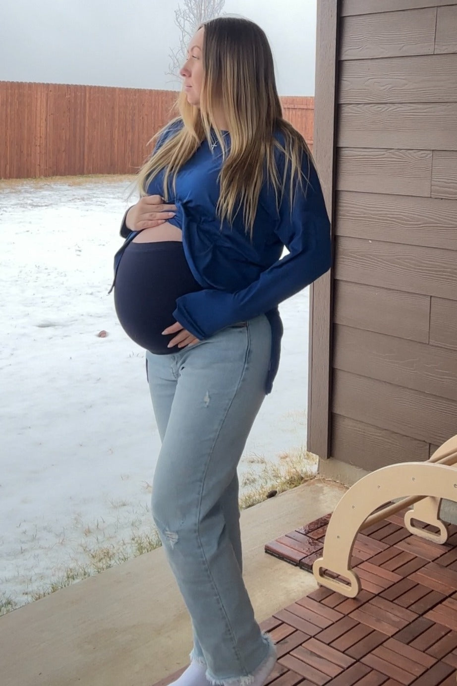 woman wearing a navy blue maternity top and light wash jeans, holding her baby bump while standing outside
