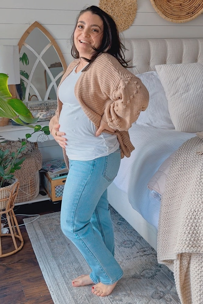 woman wearing a white maternity top and a light brown cardigan, showing her baby bump, with blue jeans, standing in a bedroom