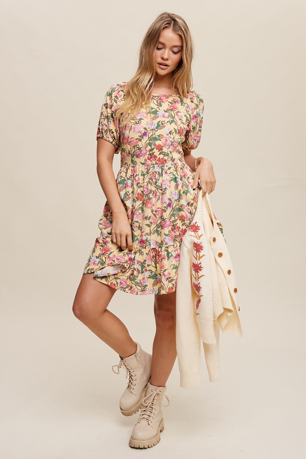 Woman wearing a yellow floral mini dress and beige combat boots holding a cream cardigan