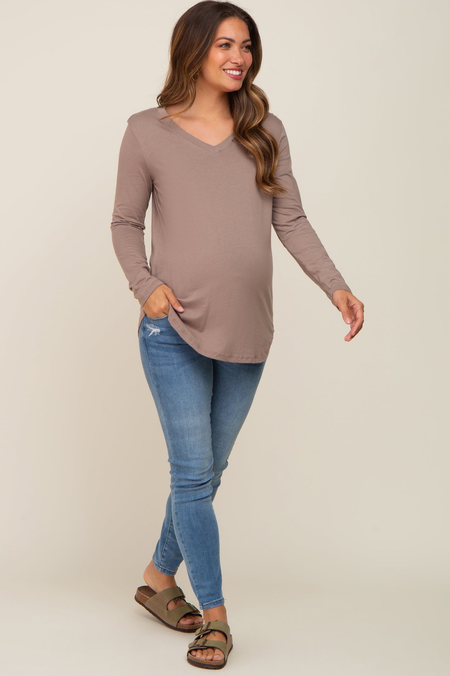 woman wearing a taupe long sleeve maternity top and blue skinny jeans with a visible baby bump