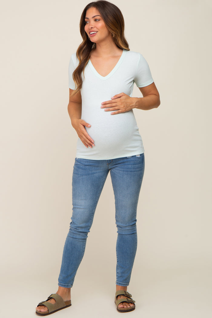woman wearing a light green v-neck maternity shirt and blue jeans, cradling her baby bump