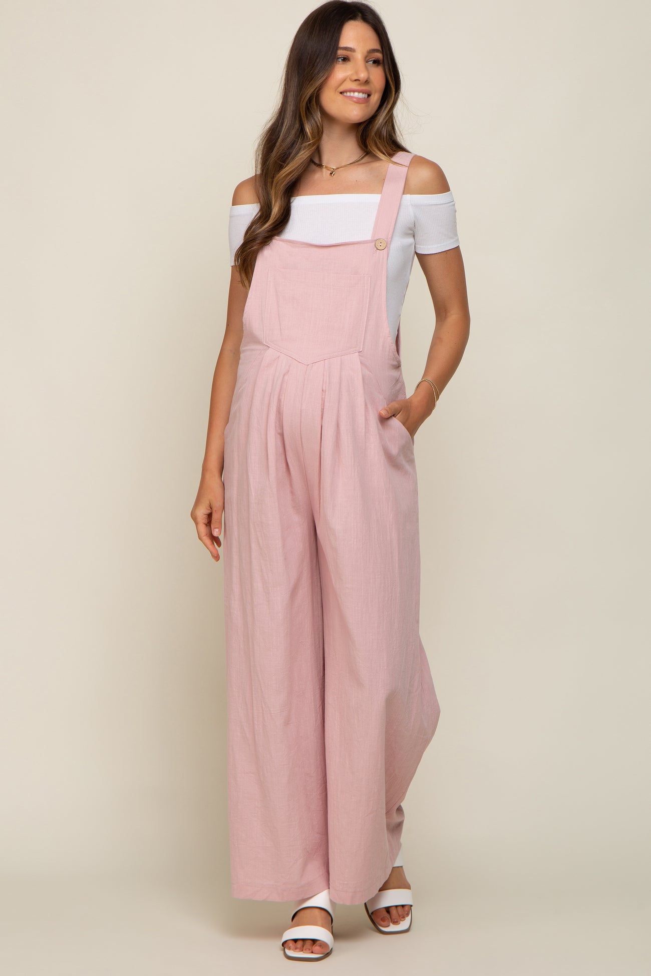 woman wearing a light pink maternity jumpsuit with off-the-shoulder white top and side pockets