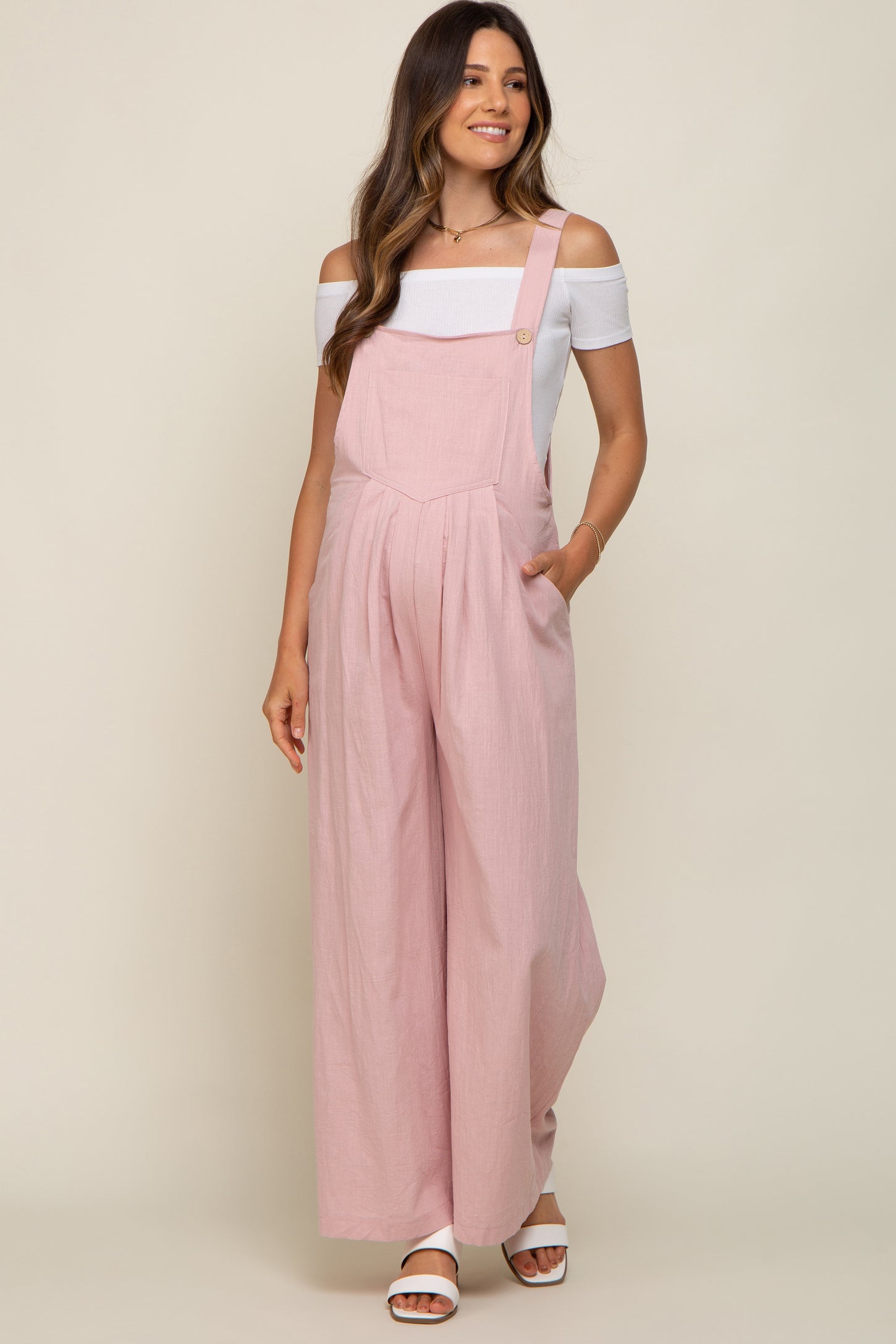 woman wearing a light pink maternity jumpsuit with off-the-shoulder white top and side pockets