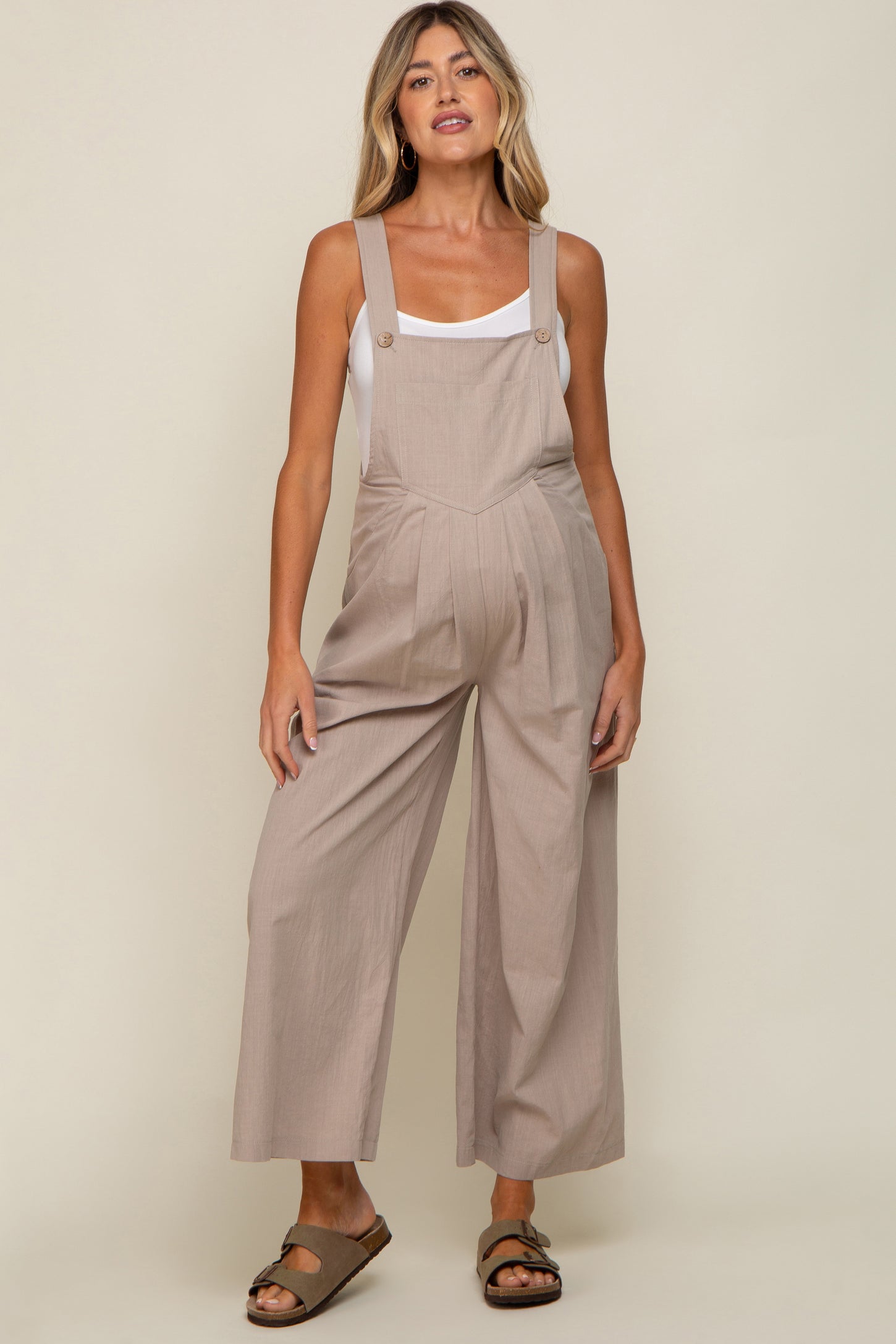 woman wearing a light brown maternity jumpsuit with wide legs and buttons, showcasing her baby bump.
