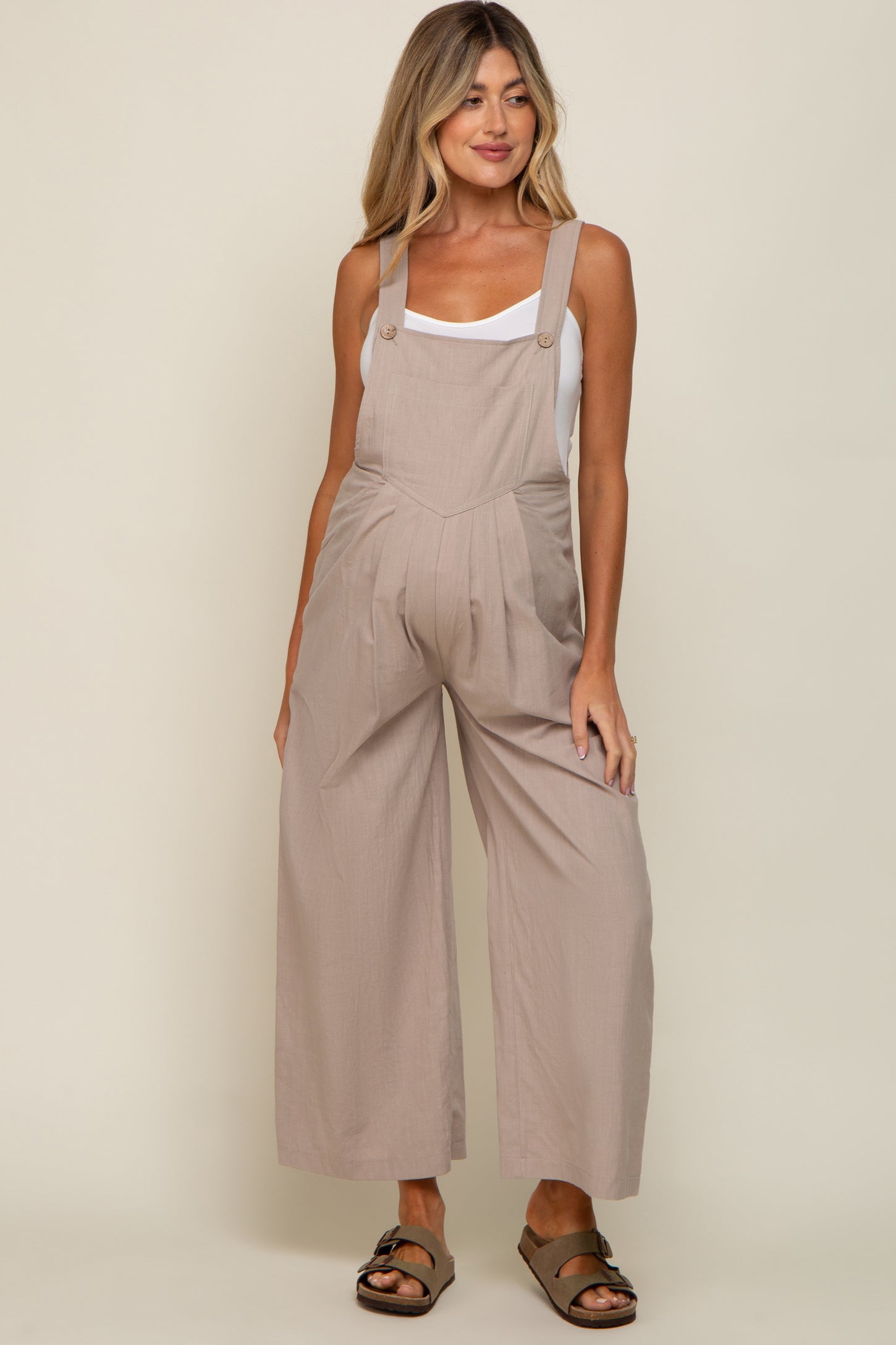 woman wearing a light beige maternity jumpsuit with wide legs and adjustable straps, showing her baby bump