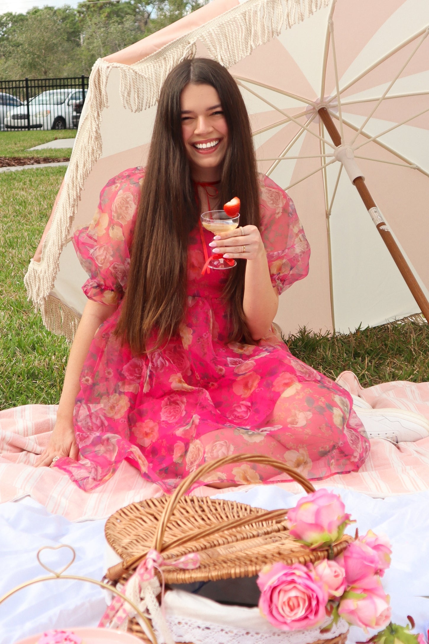 Woman in pink floral dress having a picnic with a drink under a fringed umbrella on a lawn