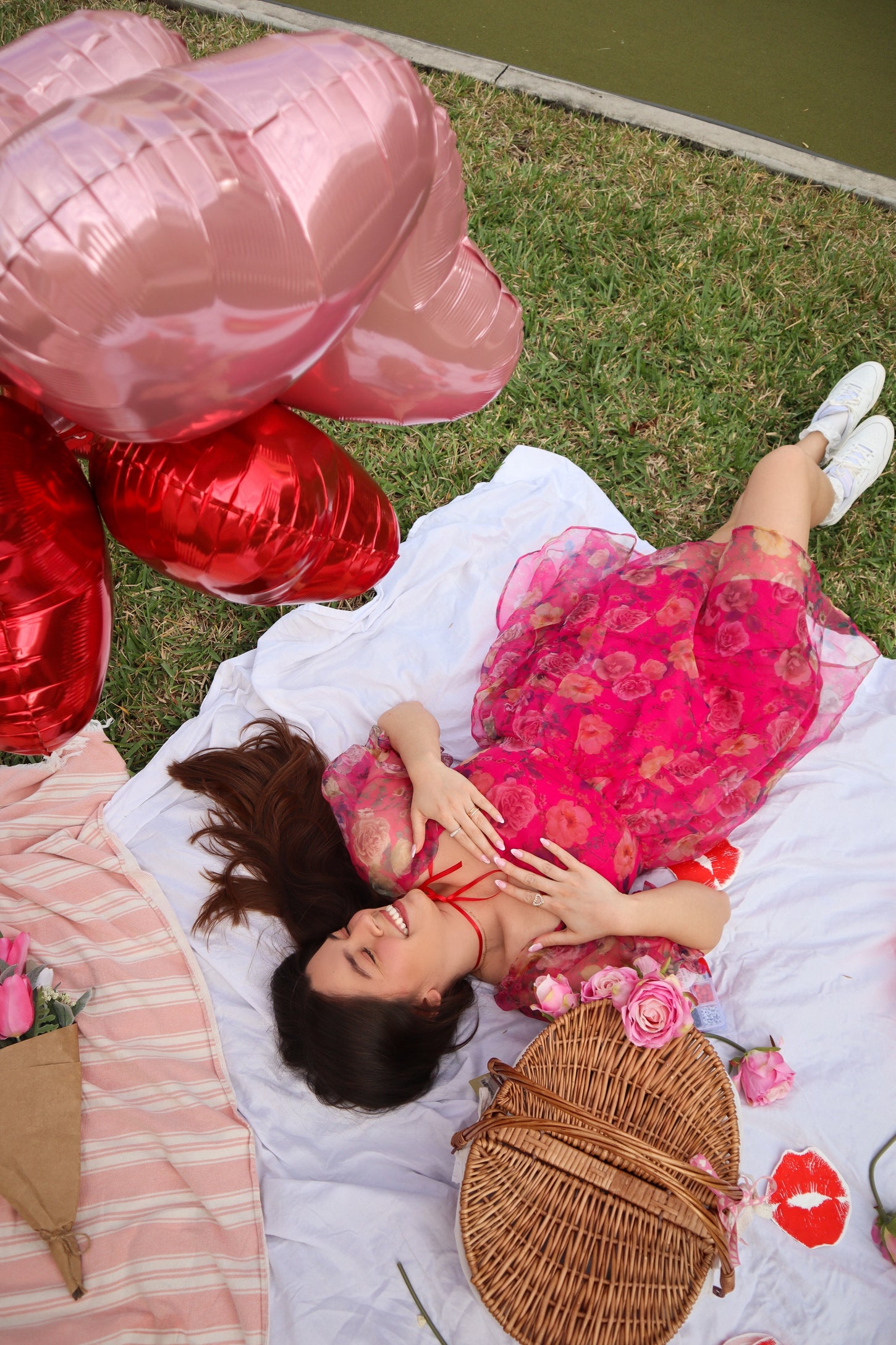 woman lying on a picnic blanket in a pink floral dress surrounded by heart-shaped balloons and a wicker basket