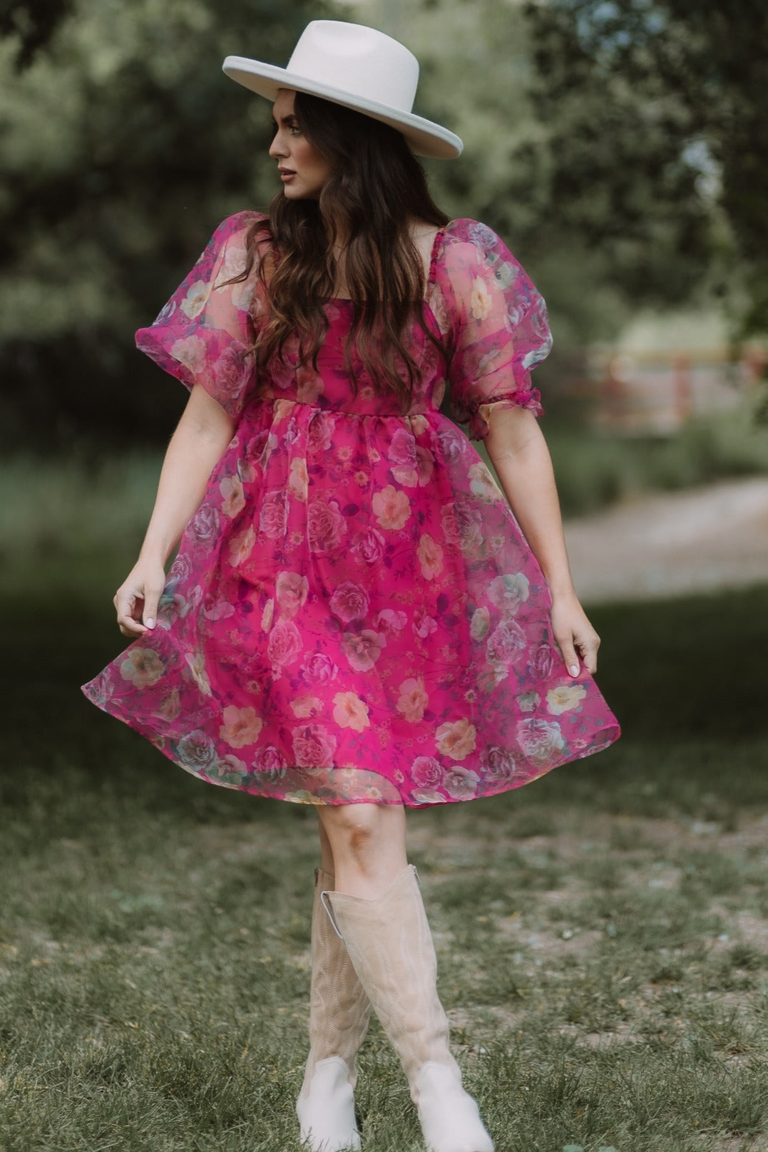 woman wearing a pink floral puff sleeve dress and a wide-brimmed white hat in an outdoor setting