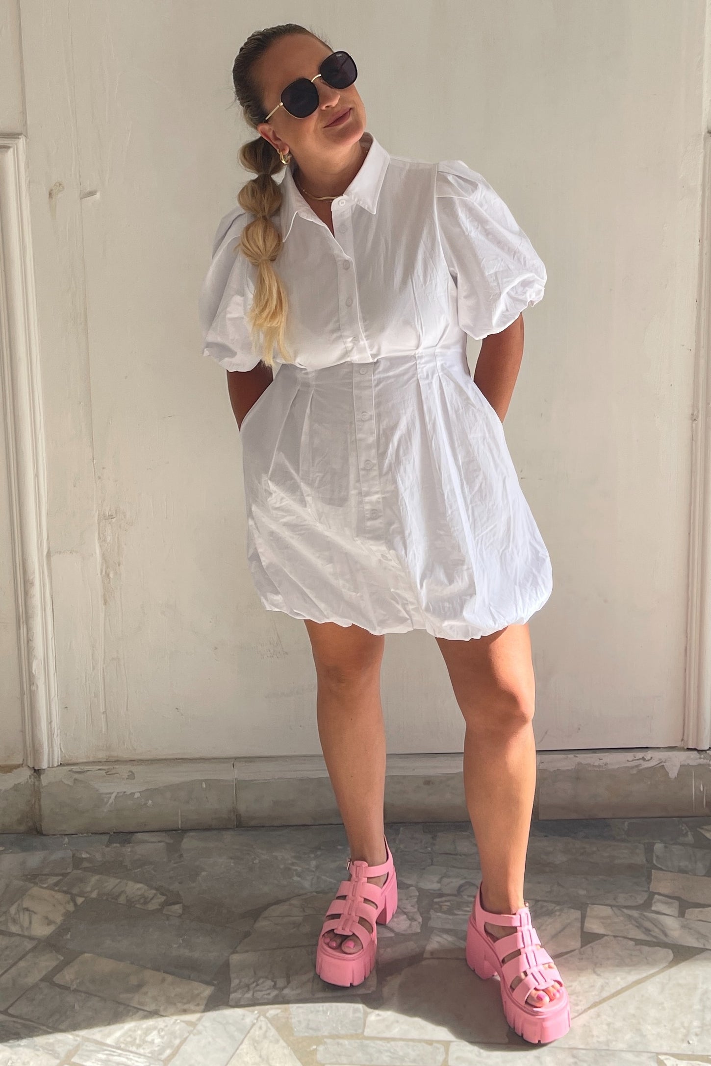 woman wearing a white puff sleeve shirt dress and pink platform sandals, standing against a white wall