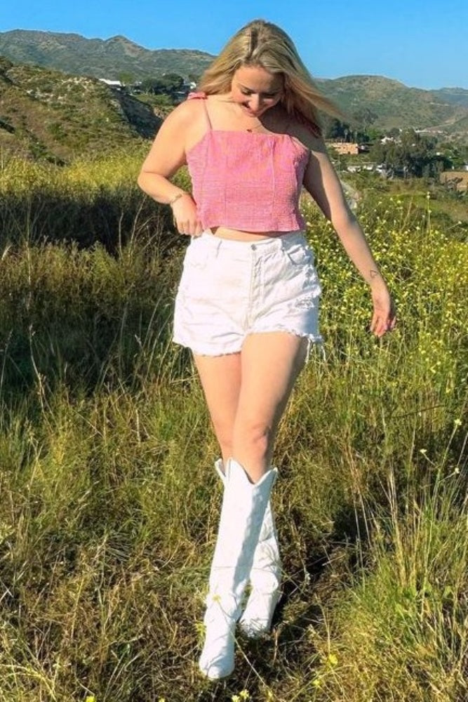 woman wearing a pink cropped top and white high-waisted shorts, paired with white knee-high boots in a scenic outdoor setting