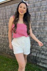 woman wearing a pink gingham crop top and white linen shorts, smiling outdoors with a headband in her hair