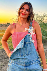 Woman wearing denim overalls and a red gingham top smiling outdoors at sunset