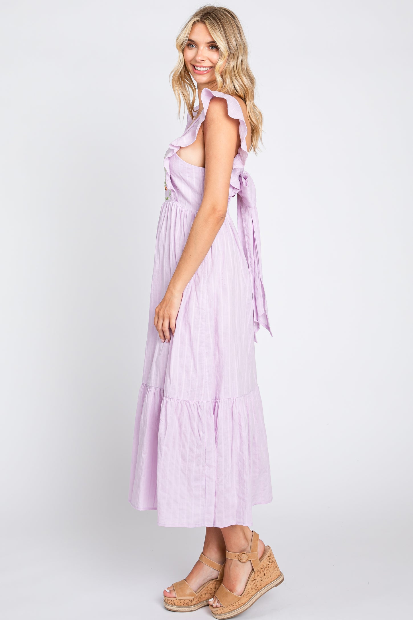 Woman wearing a lavender ruffle sleeve midi dress and tan wedge sandals against a white background
