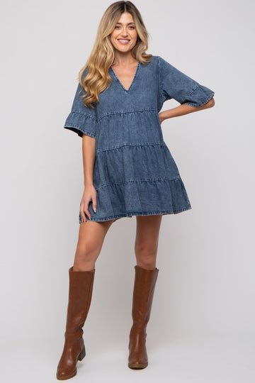 woman wearing a blue denim maternity dress with a v-neck and tiered layers, standing in brown knee-high boots
