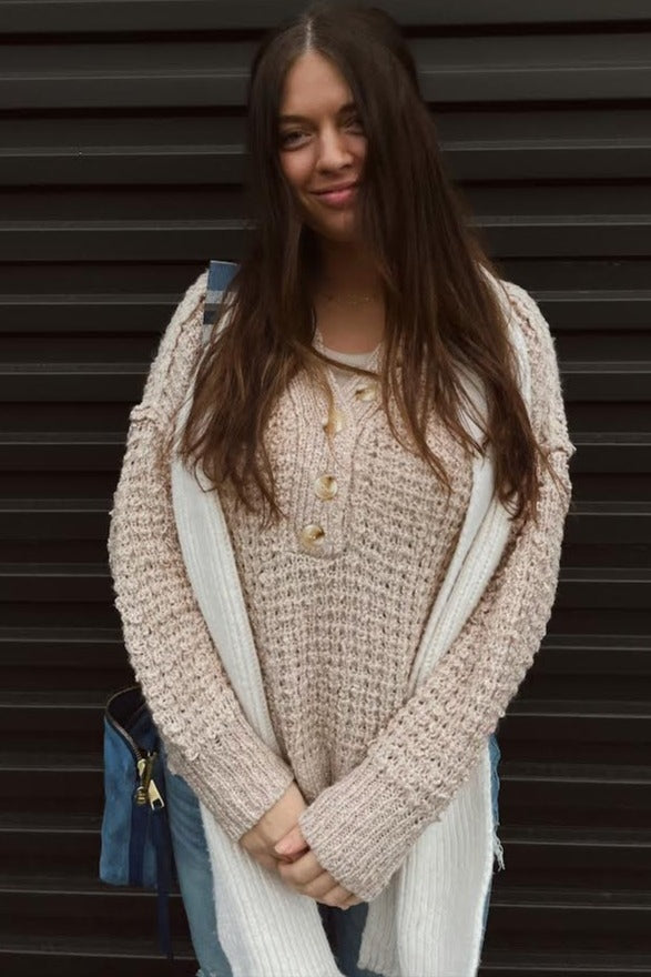 woman wearing a light beige chunky knit sweater with button details and blue jeans, posing with a smile