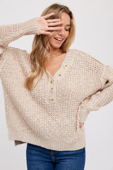 woman wearing a beige oversized knit sweater with a v-neck and button details, smiling and posing with her hand up