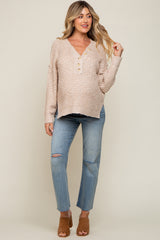 woman wearing a beige knit maternity sweater with buttons and distressed blue jeans, standing in front of a neutral background