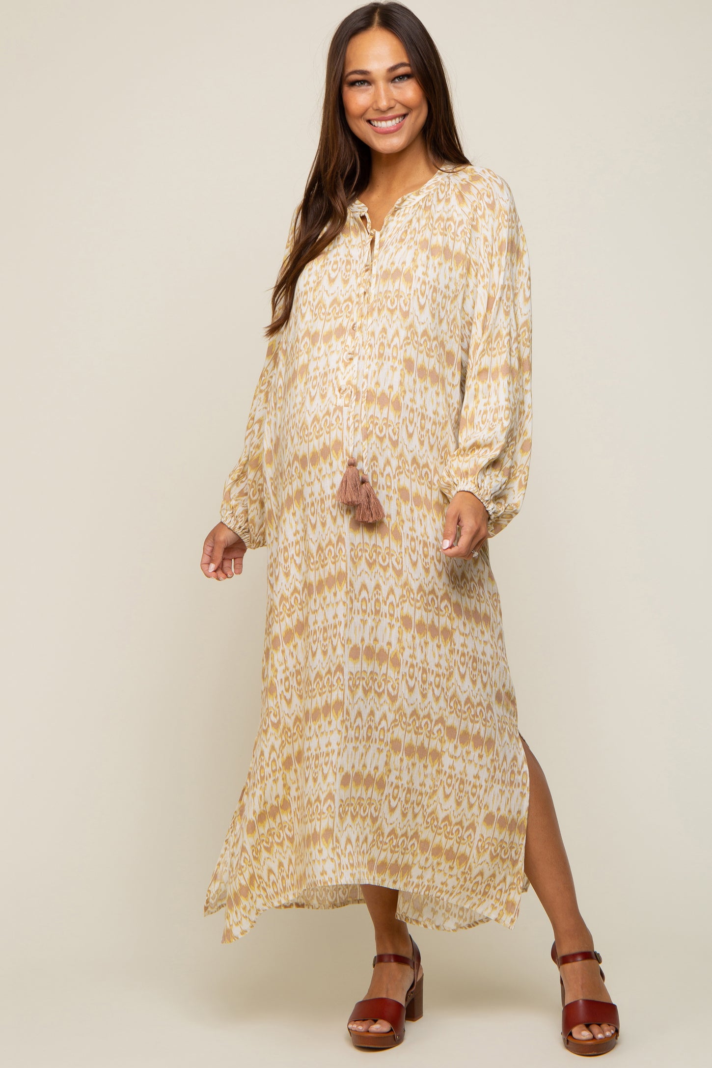 Smiling pregnant woman wearing a cream and yellow ikat print maternity maxi dress with long sleeves and tassels