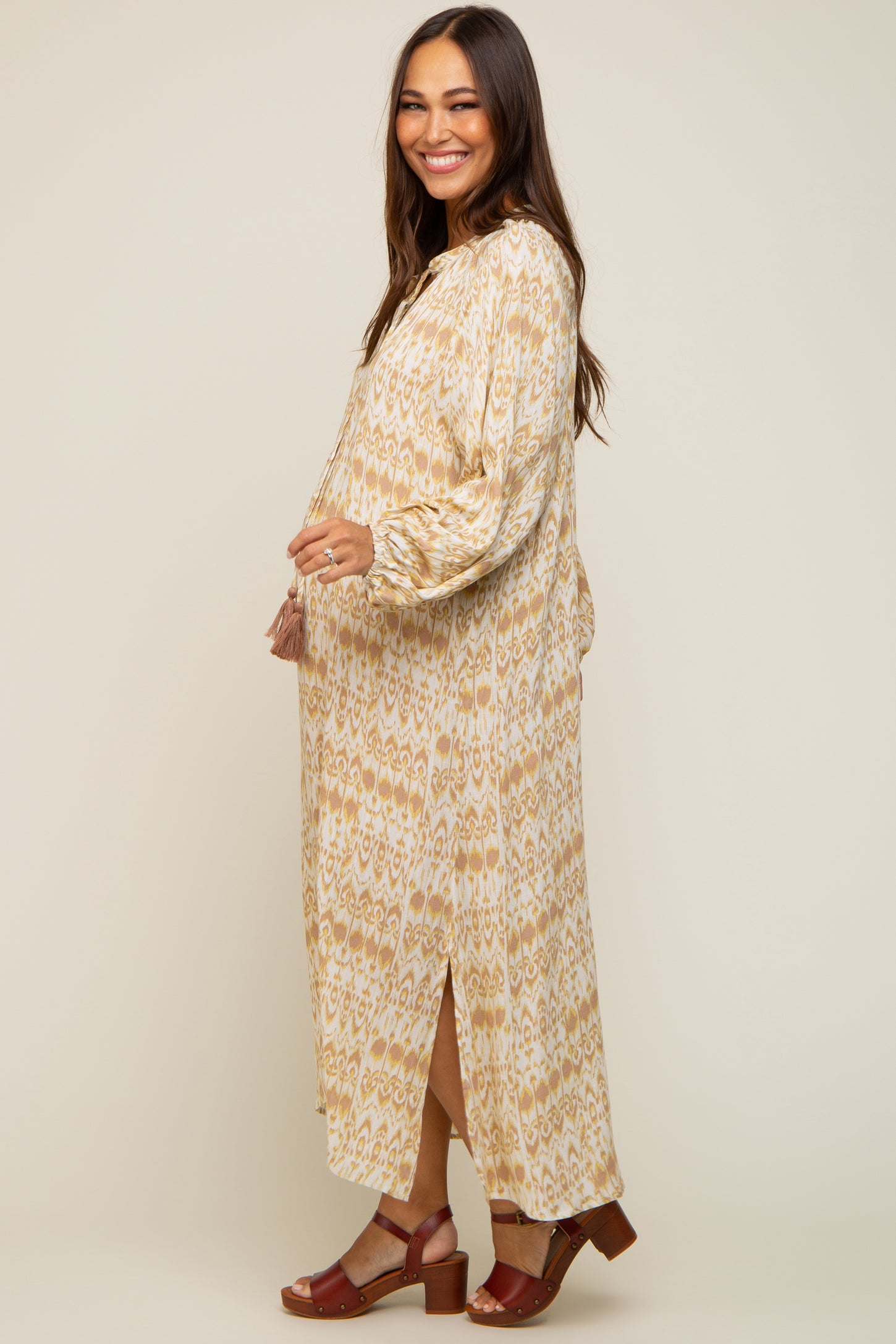 woman wearing a beige and brown patterned maternity maxi dress with a side slit and holding her baby bump