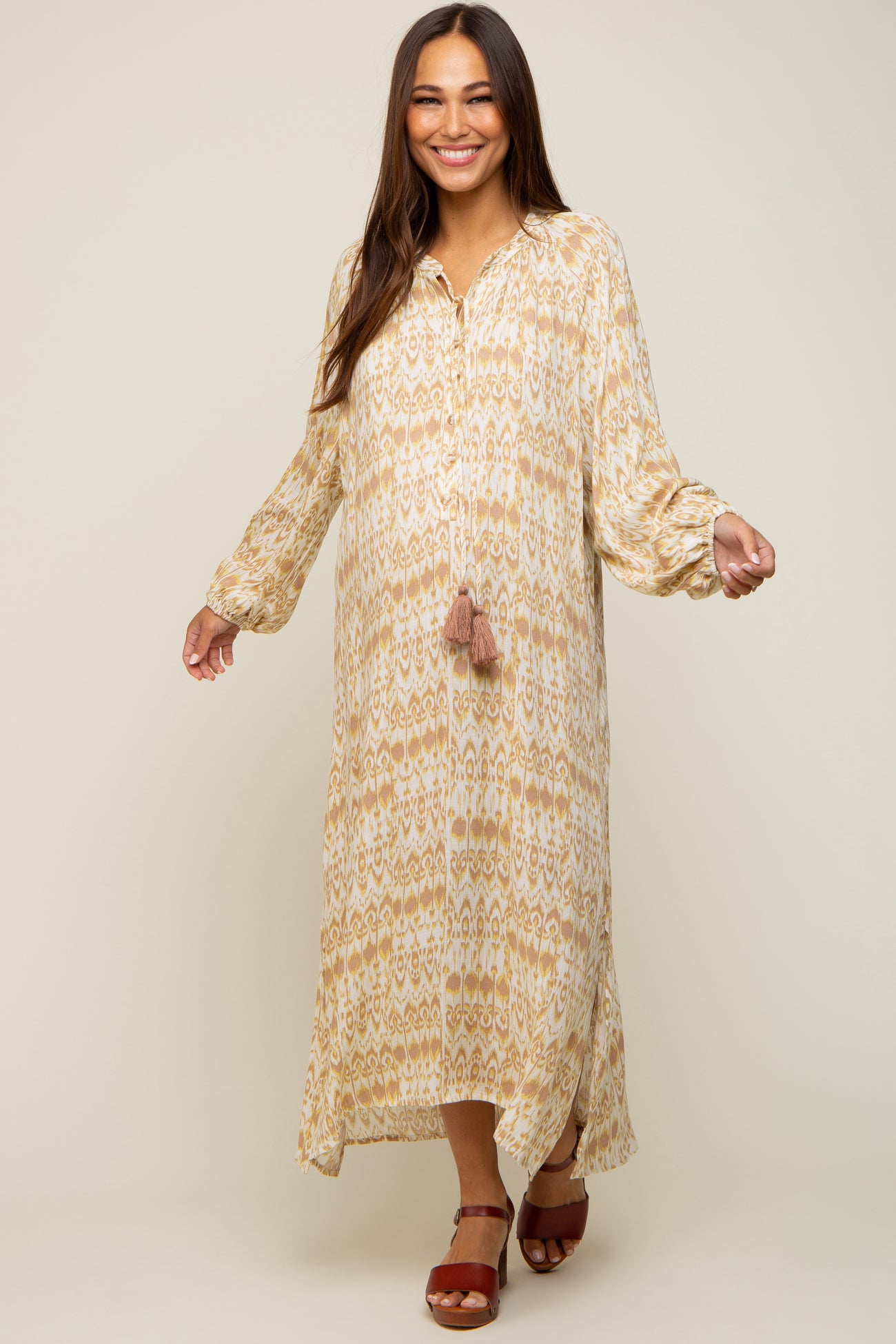 woman wearing a patterned cream and gold maternity maxi dress with long sleeves and a tassel detail