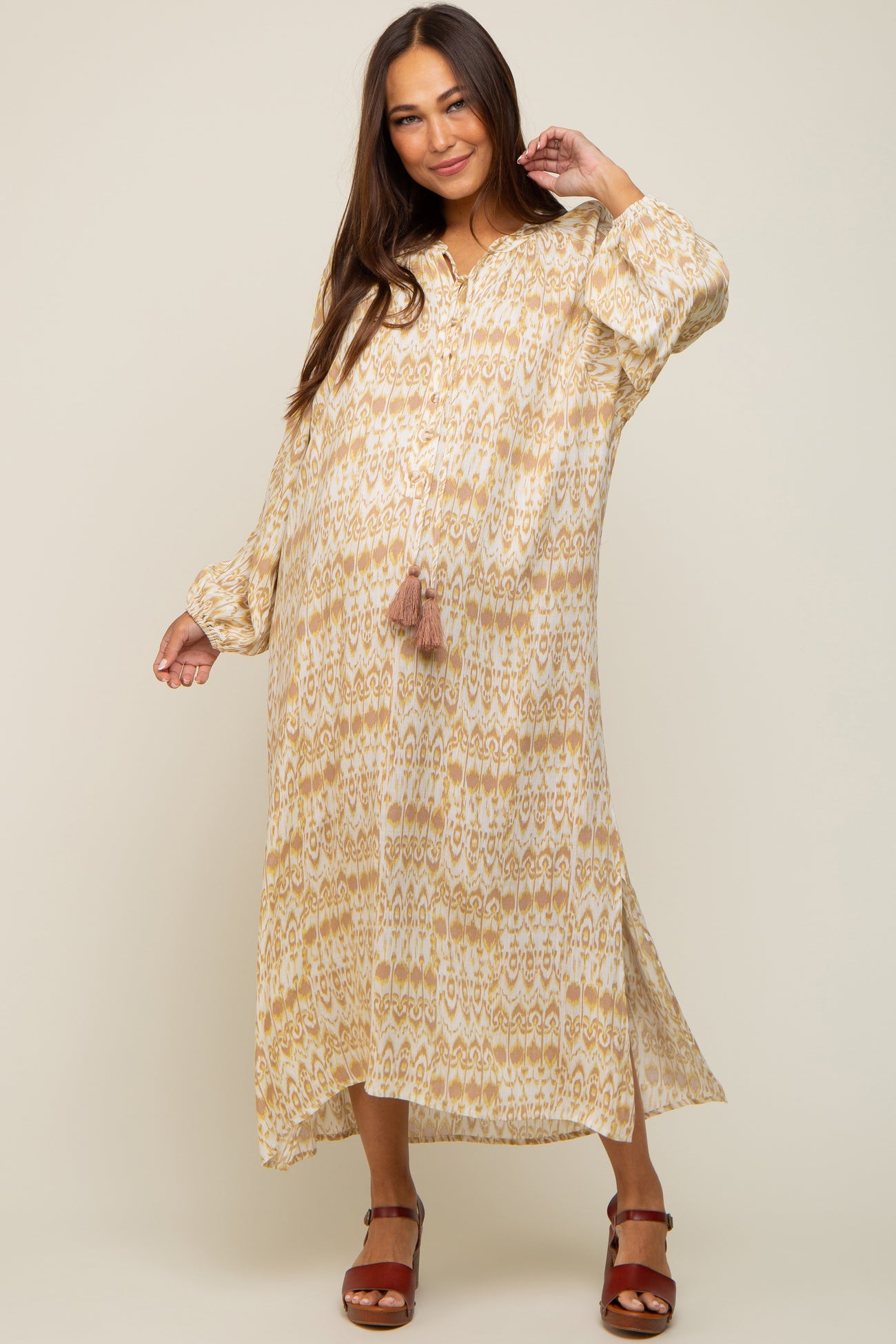 woman wearing a cream and gold patterned maternity dress with long sleeves and a tassel detail, showcasing her baby bump
