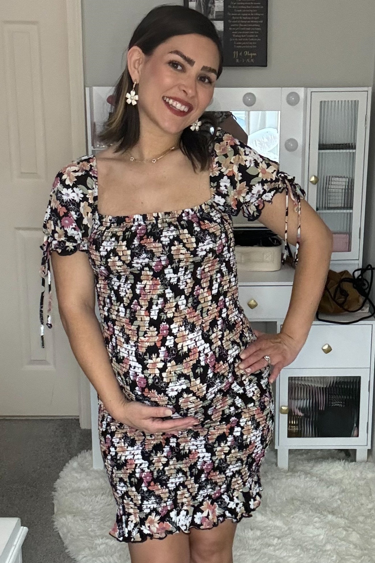 Smiling pregnant woman in a black floral smocked maternity dress cradling her baby bump in front of a white vanity