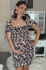 Smiling pregnant woman in a black floral smocked maternity dress cradling her baby bump in front of a white vanity