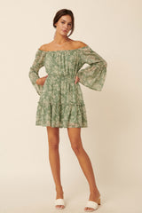 woman wearing a green floral off-shoulder dress with bell sleeves and a ruffled hem, standing against a neutral background