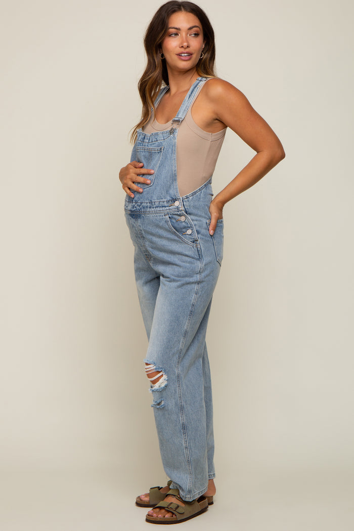 Pregnant woman wearing light wash denim maternity overalls and a beige tank top against a neutral background