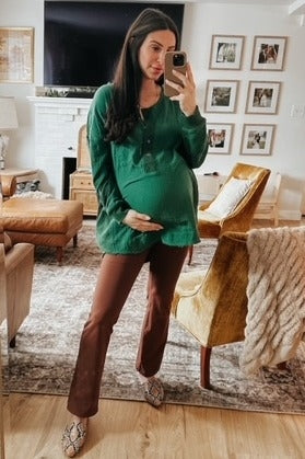 woman wearing a green maternity top and brown maternity pants, holding her baby bump in a cozy living room setting