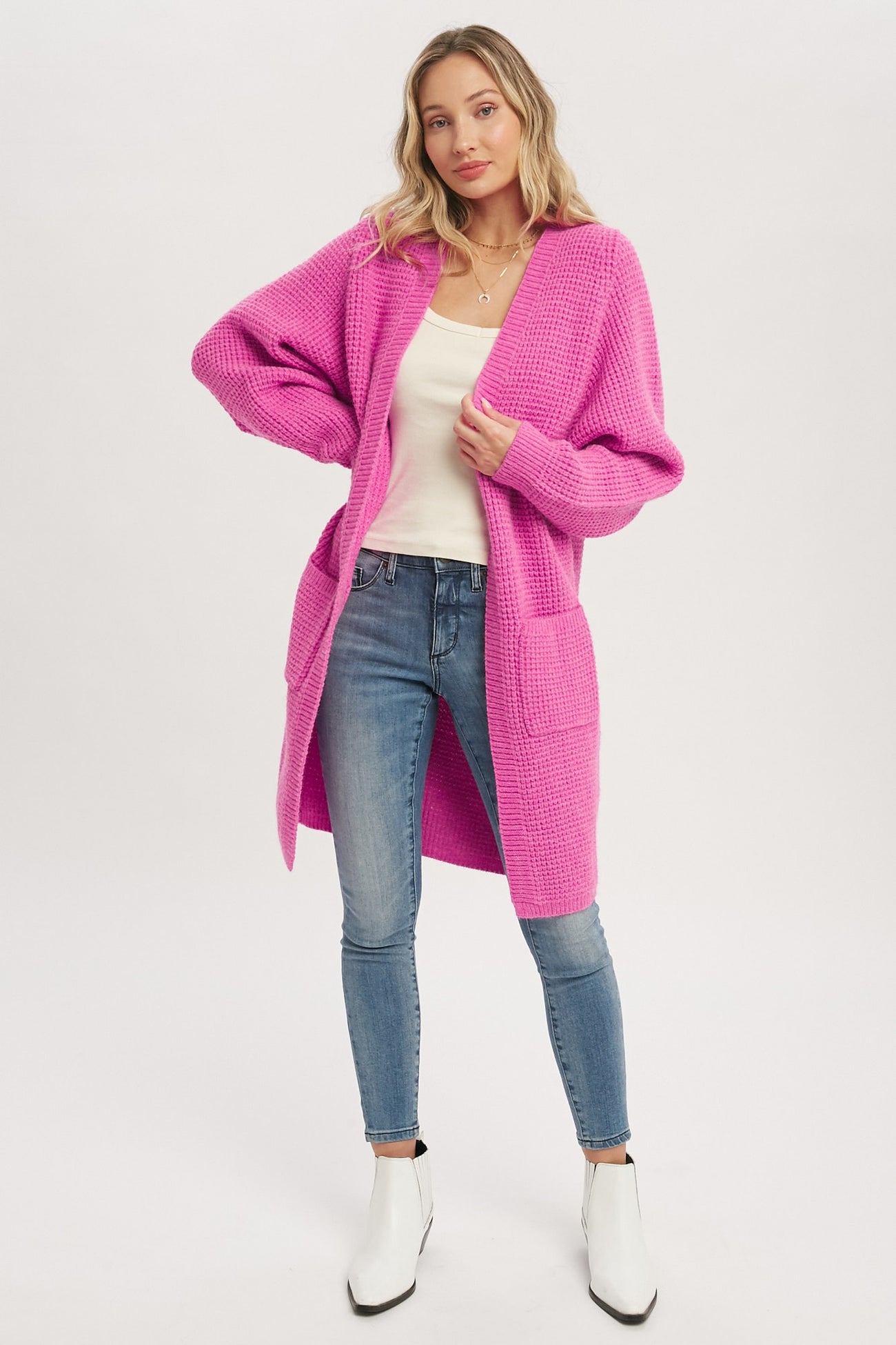 woman wearing a long bright pink waffle knit cardigan with blue skinny jeans and white ankle boots