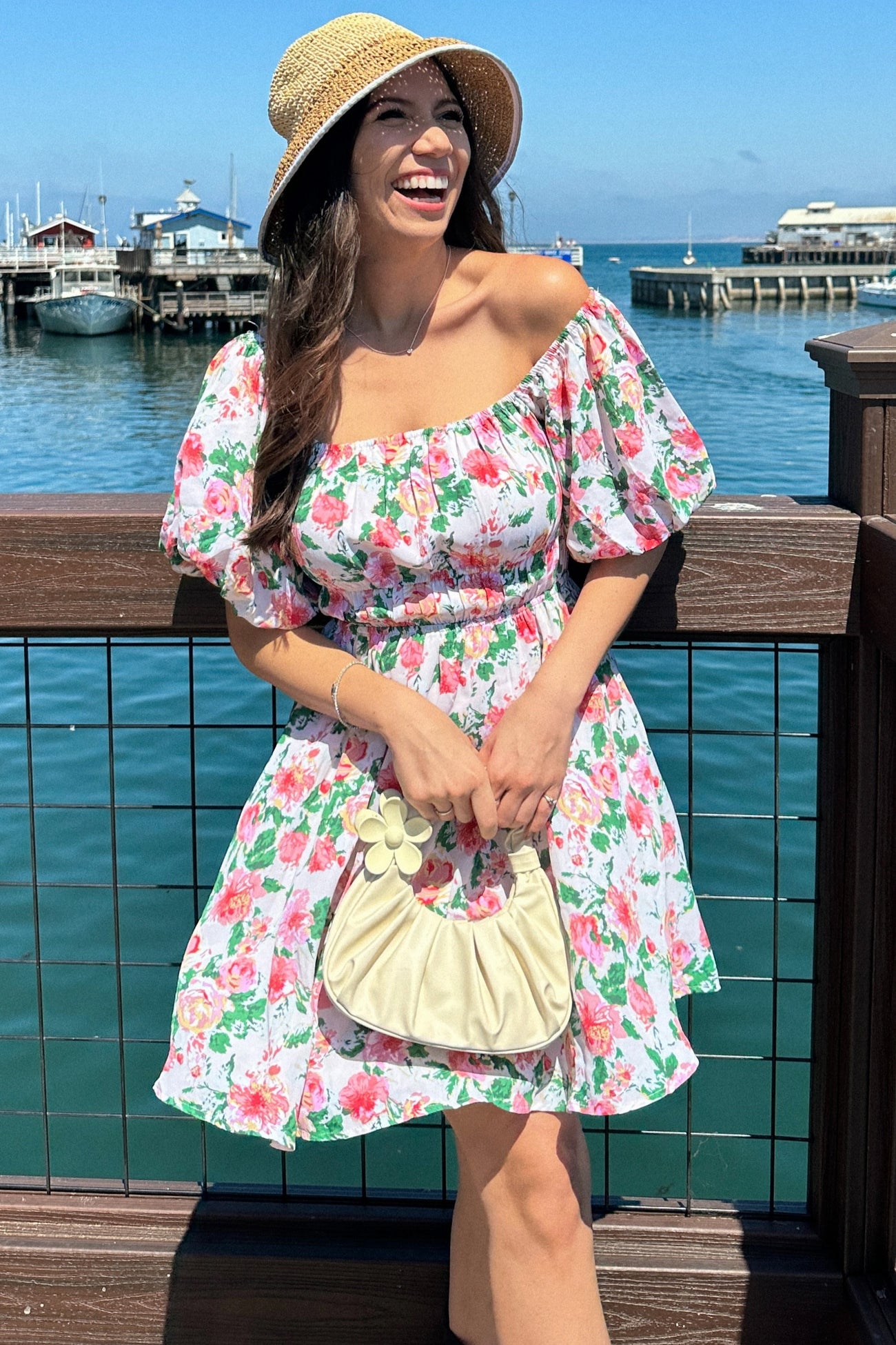 woman wearing a floral off-the-shoulder dress with puff sleeves and holding a cream handbag, smiling by the water