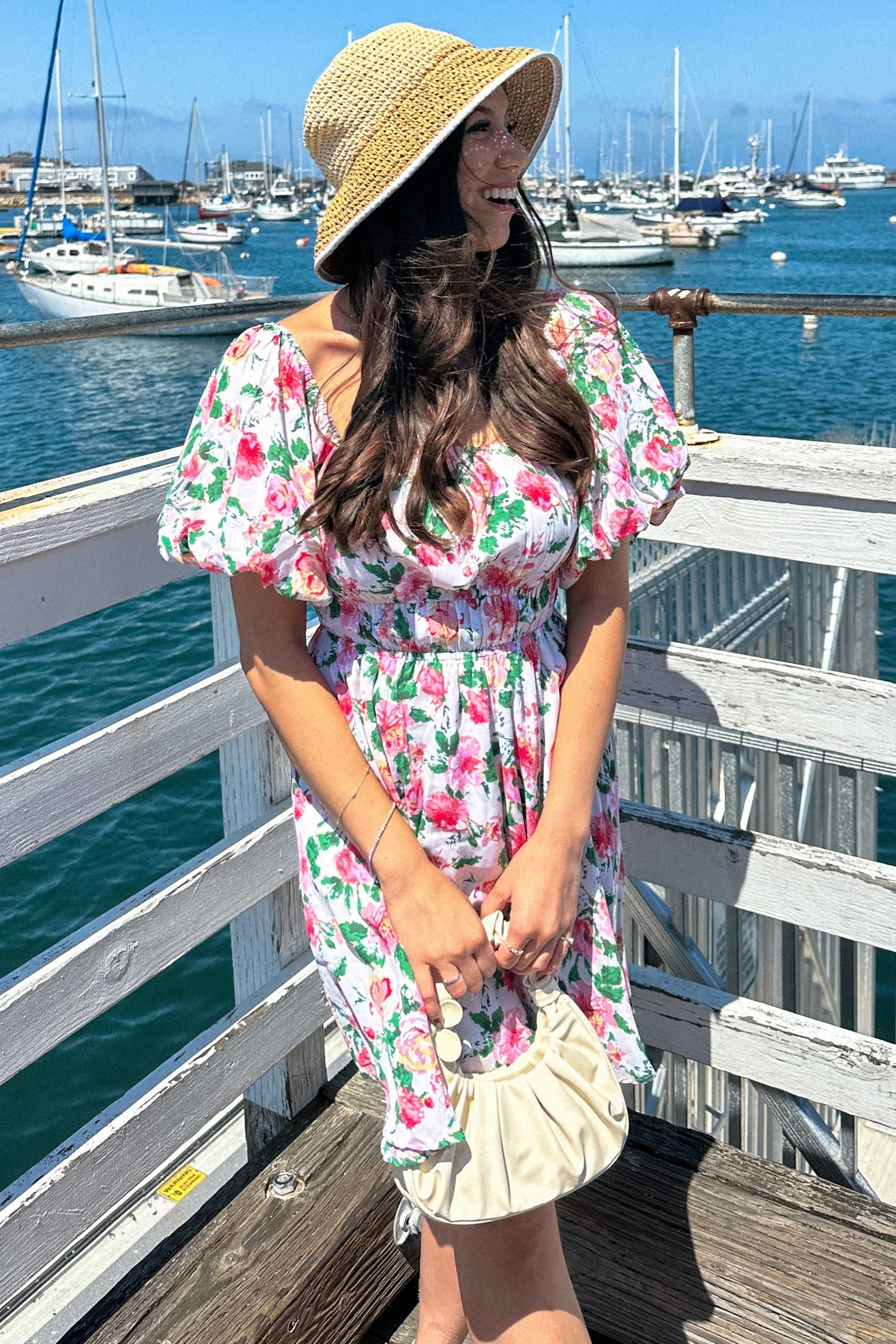 woman wearing a floral puff sleeve dress and a straw hat, standing by the water with boats in the background