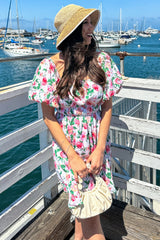 woman wearing a floral puff sleeve dress and a straw hat, standing by the water with boats in the background