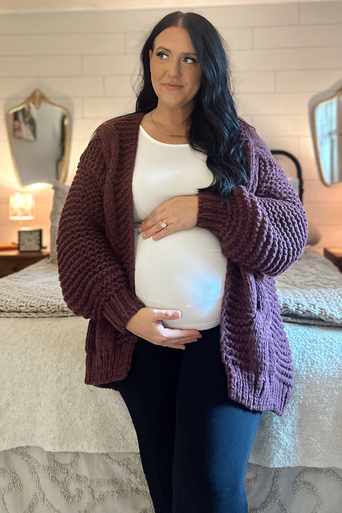 woman wearing a maroon chunky knit cardigan and white top, holding her baby bump, black pants, standing in a bedroom