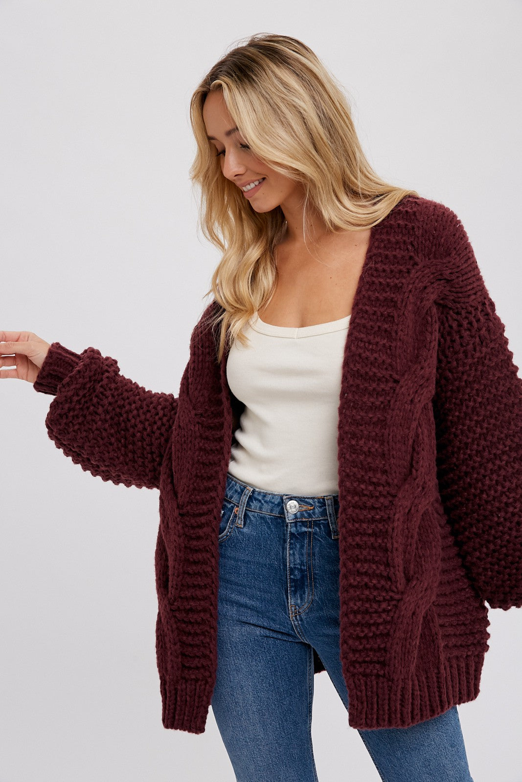 woman wearing a burgundy chunky knit cardigan with cable details over a cream tank top and blue jeans